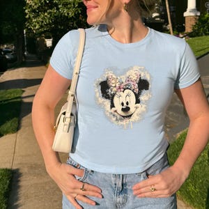 May include: Light blue t-shirt featuring a Minnie Mouse graphic. The graphic shows Minnie Mouse's face and bow, surrounded by a floral design. The shirt is paired with blue jeans. The text "COLOR: LIGHT BLUE GILDAN" is in the upper right corner.