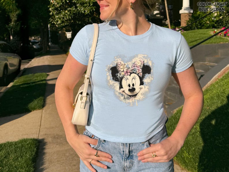May include: Light blue t-shirt featuring a Minnie Mouse graphic. The graphic shows Minnie Mouse's face and bow, surrounded by a floral design. The shirt is paired with blue jeans. The text "COLOR: LIGHT BLUE GILDAN" is in the upper right corner.