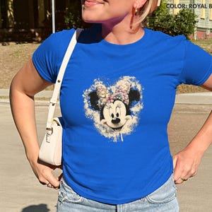 May include: Royal blue t-shirt featuring a Minnie Mouse graphic with a floral design. The graphic is centered on the shirt and includes a white and beige background. The shirt has short sleeves and a crew neck.