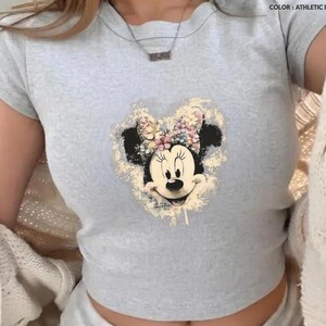 May include: A light gray t-shirt featuring a distressed Minnie Mouse graphic with a floral bow. The shirt has short sleeves and a crew neck. The text "COLOR: ATHLETIC HEATHER GILDAN" is visible in the upper right corner.