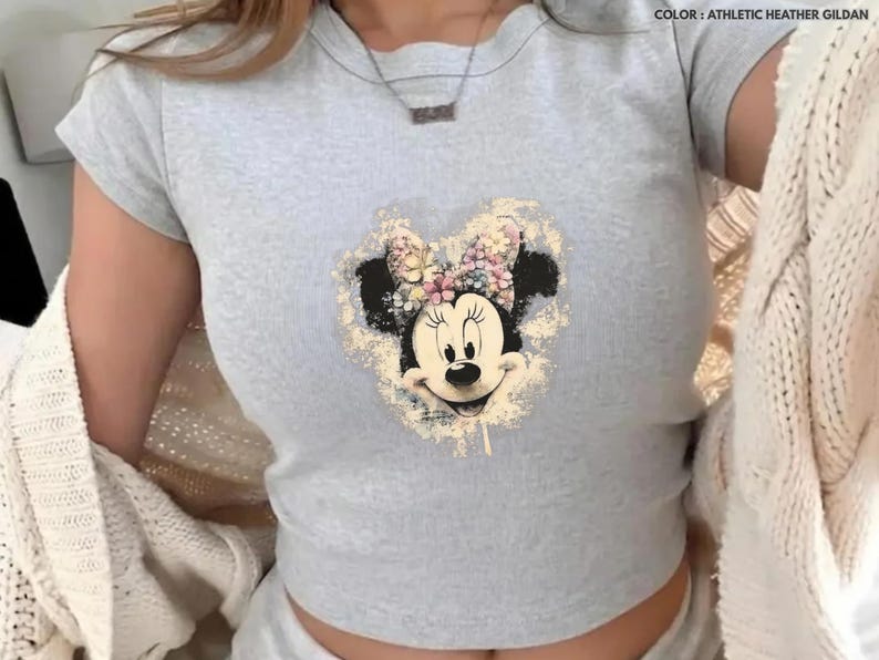 May include: A light gray t-shirt featuring a distressed Minnie Mouse graphic with a floral bow. The shirt has short sleeves and a crew neck. The text "COLOR: ATHLETIC HEATHER GILDAN" is visible in the upper right corner.