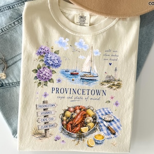 May include: An ivory t-shirt featuring a graphic print of Provincetown, Cape Cod. The design includes purple hydrangeas, sailboats on water, a lighthouse, a lobster clambake, and a wooden signpost with text reading Provincetown, Hyannis, Cape Cod, Clam Bake.