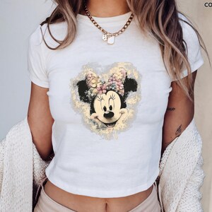 May include: White Gildan t-shirt featuring a Minnie Mouse graphic. The graphic shows Minnie Mouse's face with a floral bow and a gold-colored heart-shaped background. The shirt is short-sleeved and has a crew neck.