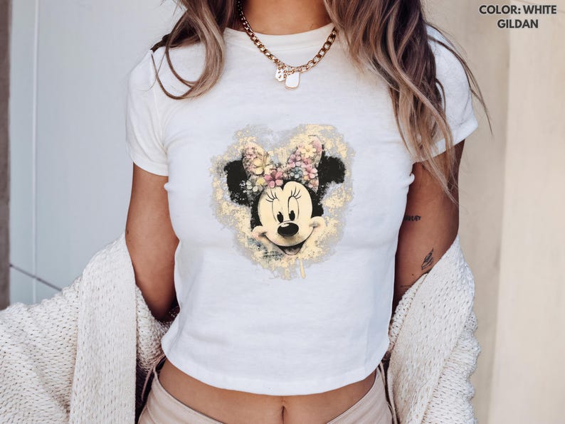 May include: White Gildan t-shirt featuring a Minnie Mouse graphic. The graphic shows Minnie Mouse's face with a floral bow and a gold-colored heart-shaped background. The shirt is short-sleeved and has a crew neck.