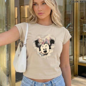 May include: A beige t-shirt featuring a Minnie Mouse graphic. The graphic is a heart-shaped design with Minnie's face and a floral bow. The shirt has short sleeves and a crew neck.