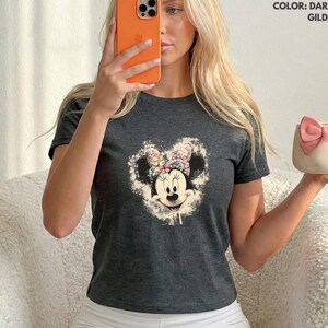 May include: Dark heather gray t-shirt featuring a Minnie Mouse graphic with a floral bow. The image also shows a white and pink mug with a bow design. The shirt is made of Gildan material.