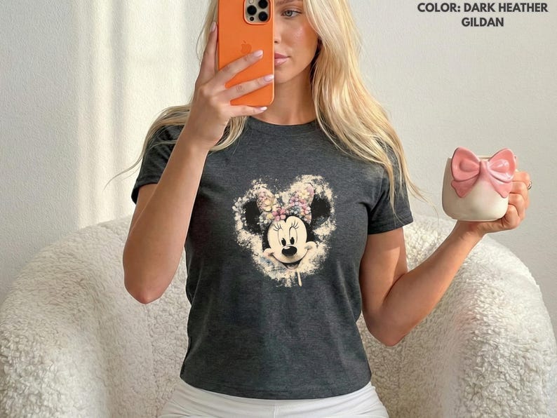 May include: Dark heather gray t-shirt featuring a Minnie Mouse graphic with a floral bow. The image also shows a white and pink mug with a bow design. The shirt is made of Gildan material.