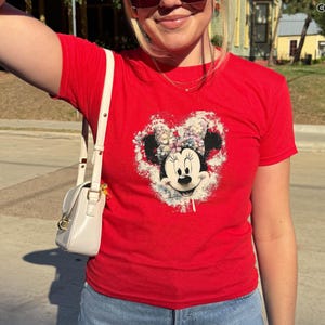 May include: A red Gildan t-shirt featuring a Minnie Mouse graphic in a heart shape. The graphic is a distressed design with a white background. The shirt has short sleeves and is worn with blue jeans and a white purse.