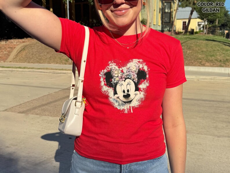 May include: A red Gildan t-shirt featuring a Minnie Mouse graphic in a heart shape. The graphic is a distressed design with a white background. The shirt has short sleeves and is worn with blue jeans and a white purse.