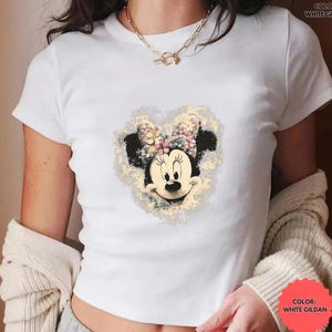 May include: White Gildan t-shirt featuring a Minnie Mouse graphic. The graphic shows Minnie's face with a floral bow and a heart-shaped background. The text "COLOR: WHITE GILDAN" is printed on the shirt.