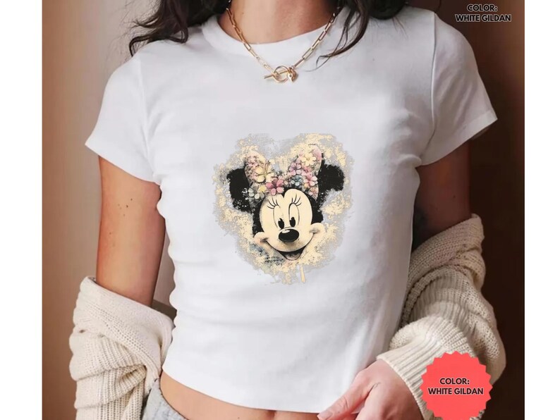 May include: White Gildan t-shirt featuring a Minnie Mouse graphic. The graphic shows Minnie's face with a floral bow and a heart-shaped background. The text "COLOR: WHITE GILDAN" is printed on the shirt.