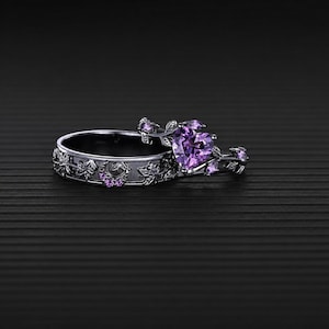 Könnte beinhalten: Ein Ring-Set aus dunkel getöntem Metall mit einem Band, das geprägte Blattmuster aufweist, und einem zweiten Ring mit einem herzförmigen violetten Edelstein als Mittelstück. Kleine violette Akzentsteine sind in das Blattdesign eingearbeitet.