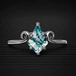 Könnte beinhalten: Ein silberfarbener Ring mit einem drachenförmigen Moosachat-Edelstein, der grüne dendritische Einschlüsse aufweist. Der Stein wird von vier Krappen gehalten und ist von polierten Metallverzierungen an beiden Seiten der Ringschiene flankiert.