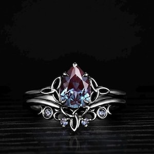 Könnte beinhalten: Ein silbernes Ring-Set mit einem birnenförmigen, farbwechselnden Edelstein. Der Ring hat ein verziertes Design mit zarten Wirbeln und kleinen, runden Akzentsteinen. Der Edelstein wechselt von tiefem Lila zu Türkis.