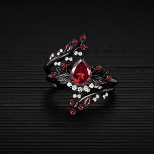 Könnte beinhalten: Schwarzer Ring mit einem großen, birnenförmigen roten Edelstein in der Mitte, umgeben von kleineren roten und weißen Edelsteinen. Der Ring hat ein florales Design mit schwarzen Metallzweigen und Blättern.