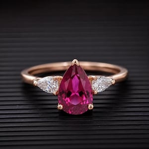 Könnte beinhalten: Ein Ring aus Roségold mit einem großen, birnenförmigen, leuchtend pinken Mittelstein, der von Krappen gehalten wird. Der Mittelstein ist auf jeder Seite von einem kleineren, birnenförmigen, klaren weißen Stein flankiert. Die Ringschiene ist glatt.