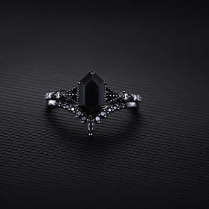 Könnte beinhalten: Ein schwarzer Edelsteinring mit geometrischem Schliff, gefasst in einem dunklen Metallband. Der Ring hat einen zentralen, großen schwarzen Stein und ist mit kleineren schwarzen Steinen akzentuiert. Das Design ist aufwendig und detailliert, mit einer Vintage- oder Gothic-Ästhetik.