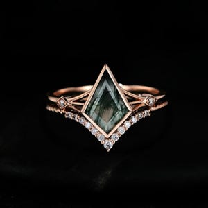 Könnte beinhalten: Ein Ring-Set aus Roségold mit einem drachenförmigen Moosachat-Mittelstein mit dunkelgrünen Einschlüssen. Die Ringschiene ist mit kleinen klaren Steinen besetzt, ergänzt durch einen V-förmigen Vorsteckring mit runden, klaren Steinen.