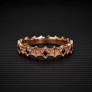 Könnte beinhalten: Ein Ring aus Roségold mit einem strukturierten, gezackten Band und einer Reihe von gleichmäßig verteilten, schwarzen rautenförmigen Steinen. Das Band hat ein sich wiederholendes architektonisches Muster mit spitzen Kanten.