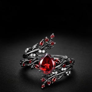 Könnte beinhalten: Ein schwarzer Ring mit einem großen, tropfenförmigen roten Edelstein. Der Ring hat ein Blatt- und Beeren-Design mit kleineren roten Edelsteinen und weißen Akzenten. Der Ring ist vor einem schwarzen Hintergrund platziert.