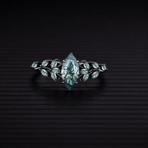 Könnte beinhalten: Ein Ring aus schwarzem Metall mit einem zentralen, marquise-geschliffenen Moosachat mit dunkelgrünen Einschlüssen. Das Band ist mit einer Reihe kleiner, blattförmiger Moosachat-Akzente verziert, die sich vom Mittelstein zu den Seiten erstrecken.