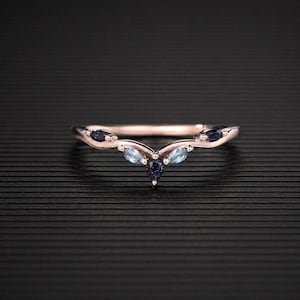 Könnte beinhalten: Ein roségoldener Ring mit geschwungenem Design. Der Ring hat drei hellblaue, marquiseförmige Edelsteine und drei dunkelblaue, runde Edelsteine. Der Ring ist vor einem schwarzen Hintergrund platziert.