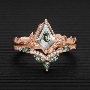 Könnte beinhalten: Ein Ring-Set aus Roségold mit einem drachenförmigen Moosachat-Mittelstein mit grünen Einschlüssen. Die Ringschiene hat ein blattartiges Design und der passende chevronförmige Ring ist mit kleinen weißen Steinen und marquise-geschliffenem Moosachat besetzt.