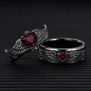 Könnte beinhalten: Ein Set aus zwei dunkelgrauen Metallringen. Ein Ring zeigt einen markanten herzförmigen roten Edelstein, der von detaillierten Flügeln flankiert wird. Der zweite Ring ist ein Band mit einem kleineren roten Herzstein und Flügelmotiven.