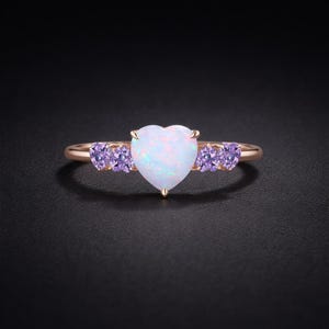 Könnte beinhalten: Ein roségoldener Ring mit einem herzförmigen weißen Opal-Mittelstein, flankiert von zwei Clustern aus kleinen, herzförmigen lila Edelsteinen. Der Ring ist vor einem dunklen Hintergrund platziert, der das Design des Schmucks hervorhebt.