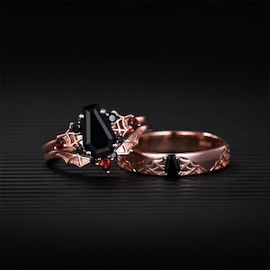 Könnte beinhalten: Roségoldenes Ring-Set mit einem Sarg-förmigen schwarzen Edelstein als Mittelstein, umgeben von kleineren schwarzen und roten Steinen. Das Band hat ein Fledermausflügel-Design. Der zweite Ring hat einen schwarzen Edelstein und ein dekoratives Band.