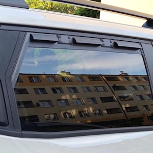 May include: A white vehicle side window featuring a black aftermarket ventilation insert installed at the top. The insert includes three small, protruding air vents along its horizontal frame, integrated into the dark window trim and roof rail.