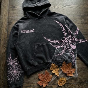 Peut inclure: Un sweat à capuche gris anthracite délavé à l'acide avec un motif rose représentant une araignée stylisée et une toile sur le devant et la manche. Le texte "YOUBERAND" est imprimé en police gothique sur la poitrine droite. Capuche et poche.