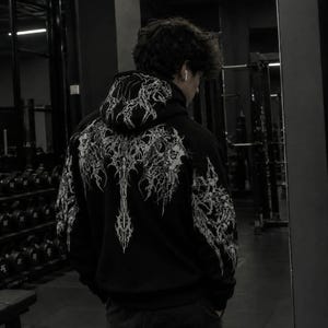 May include: A black hooded sweatshirt featuring an intricate, white, symmetrical graphic print that extends across the back and onto the sleeves. The design consists of sharp, jagged, and flowing lines that create a complex, abstract pattern.