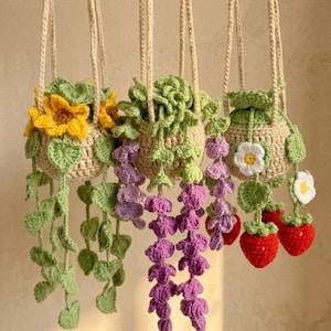 May include: Three hanging crochet plant baskets made of beige yarn. The first features yellow sunflowers and green leaves. The second has cascading purple flowers and green foliage. The third displays white flowers and red strawberries with green leaves.