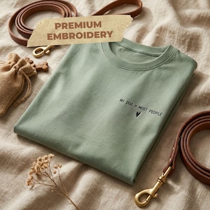 Könnte beinhalten: Ein salbeigrünes T-Shirt mit Rundhalsausschnitt und schwarzer Stickerei mit dem Text MY DOG > MOST PEOPLE sowie einem kleinen Herzsymbol darunter. Das Shirt liegt neben braunen Hundeleinen aus Leder und einem kleinen Jutebeutel mit Kordelzug.