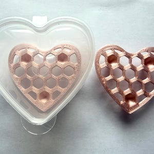 May include: A clear, heart-shaped plastic container holding a rose gold, heart-shaped insert with a honeycomb pattern, alongside a matching rose gold, heart-shaped honeycomb insert. Both inserts feature a textured, metallic finish.