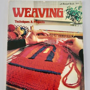 May include: The cover of a book titled "WEAVING" in red letters, with the subtitle "Techniques & Projects." The image shows a person weaving a red textile with blue, orange, and yellow accents. A basket of yarn and a small plant are also visible.