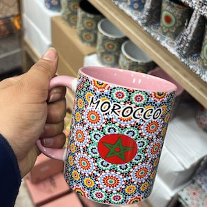Op de afbeelding: Een keramische mok met een roze handvat en binnenkant, met een kleurrijk mozaïekpatroon en het woord "MOROCCO". Het ontwerp bevat een rode cirkel met een groene ster, omringd door bekers met een vergelijkbaar patroon.
