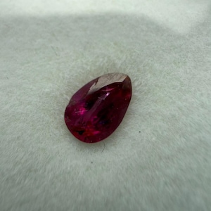 Peut inclure: Une pierre précieuse en forme de goutte d'eau, de couleur rouge profond. La gemme est posée sur un fond blanc texturé, mettant en valeur ses surfaces polies et sa clarté interne. La couleur de la pierre varie du cramoisi vif au rouge plus foncé et saturé.