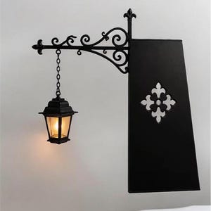 May include: A black metal wall-mounted sign bracket featuring an ornate scrollwork arm and a hanging lantern with a warm glowing light. A rectangular black sign panel is attached to the bracket, displaying a centered white quatrefoil cutout.