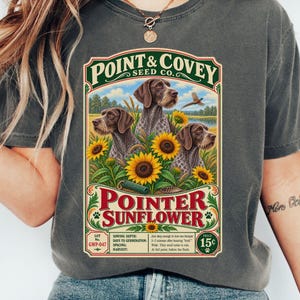 May include: A charcoal gray t-shirt featuring a vintage-style seed packet graphic. The design displays three brown and white pointer dogs surrounded by bright yellow sunflowers, with the text Point & Covey Seed Co. and Pointer Sunflower.