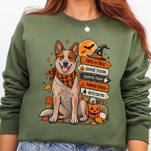 May include: An olive green crewneck sweatshirt featuring a graphic of a red heeler dog wearing an orange and black plaid bandana. The design includes a stack of directional signs labeled Trick or Treat, Hayride Station, Haunted Train, Pumpkin Express, and Witch's Flight Path, alongside a jack-o'-lantern, autumn leaves, and a witch hat.