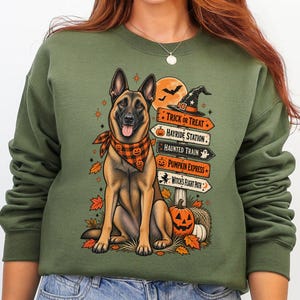 May include: An olive green crewneck sweatshirt featuring a graphic of a Belgian Malinois dog wearing an orange and black plaid bandana. The design includes a signpost with text reading Trick or Treat, Hayride Station, Haunted Train, Pumpkin Express, and Witch's Flight Path, surrounded by autumn leaves, a pumpkin, and a witch hat.