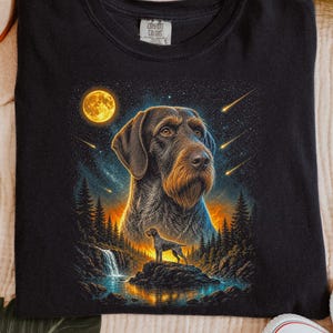 May include: A black t-shirt featuring a graphic print of a German Wirehaired Pointer dog. The design includes a large portrait of the dog above a smaller silhouette standing on a rock near a waterfall, set against a starry night sky with a full moon.