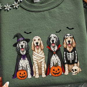 May include: An olive green sweatshirt featuring a graphic of four dogs dressed in Halloween costumes. The dogs are depicted as a witch, a mummy, a vampire, and a skeleton, accompanied by two jack-o'-lanterns, a skull, and three black bats.