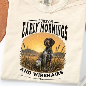 May include: A cream-colored t-shirt featuring a graphic of a German Shorthaired Pointer sitting in a field of tall grass at sunrise. The text reads BUILT ON EARLY MORNINGS AND WIREHAIRS. The shirt has a Comfort Colors brand label at the neck.
