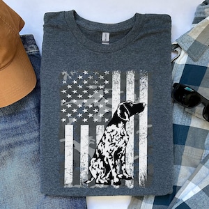 May include: Dark gray t-shirt featuring a distressed American flag design with a dog silhouette. The flag incorporates camouflage elements. A brown baseball cap with a paw print and blue jeans are also in the image.