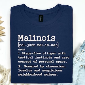 May include: A navy blue t-shirt featuring white text that defines the word Malinois as a stage-five clinger with tactical instincts, zero concept of personal space, and a personality powered by obsession, loyalty, and suspicious neighborhood noises.
