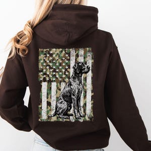 May include: A dark brown hoodie featuring a graphic of a black dog sitting in front of a camouflage American flag. The flag design incorporates white stripes and a green, brown, and white camouflage pattern.