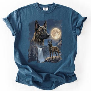 May include: A blue t-shirt featuring a detailed illustration of a dog with a waterfall, full moon, and forest scene. The dog is depicted in two sizes, with a close-up of the head and a full body shot. The design is centered on the shirt.